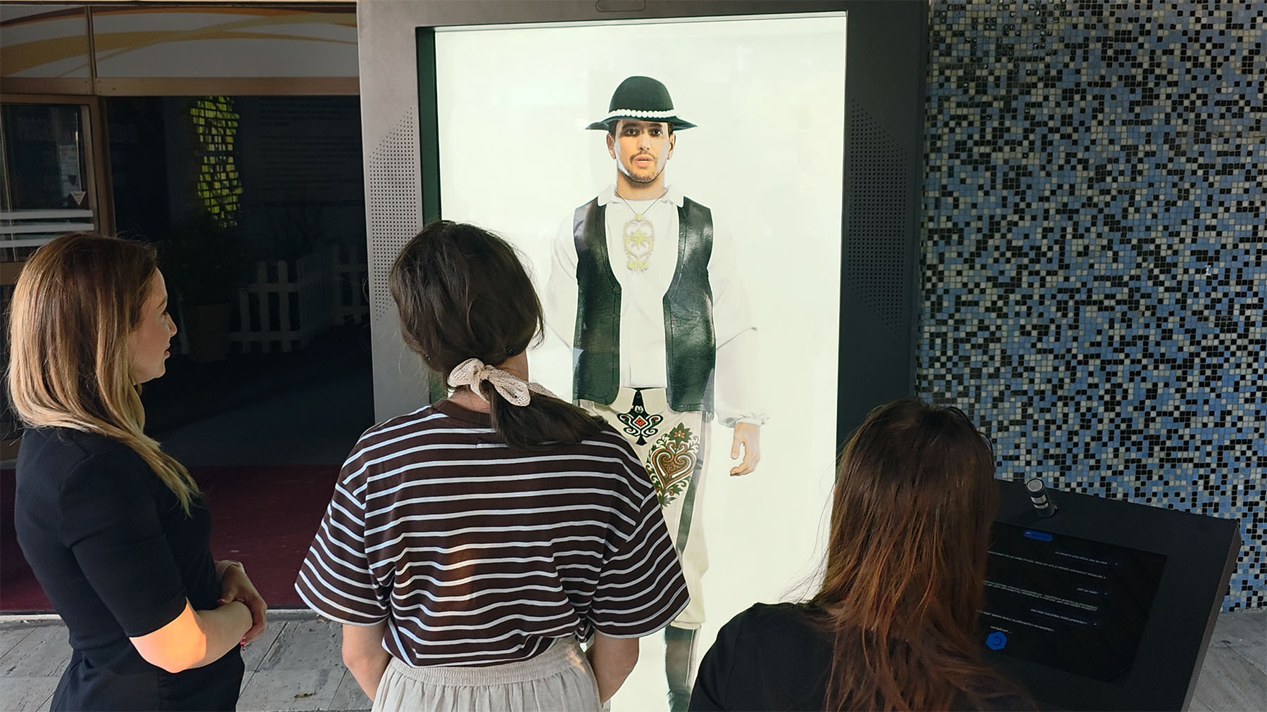 Three women stand before a large digital screen showing a man in folk attire and black hat; a control panel is to the right.
