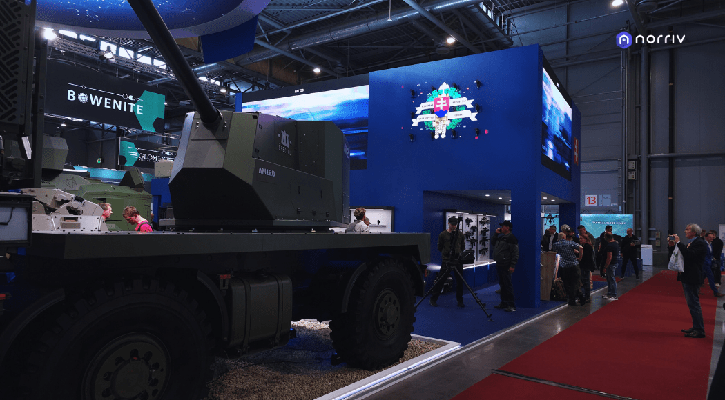 A military vehicle is displayed at an indoor exhibition. Several people observe the vehicle and other displays. A blue booth with a digital Slovak flag and norriv logo is visible in the background.