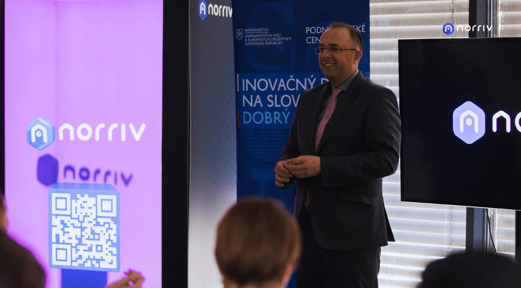 A man in a suit stands and smiles while presenting at an event. In the foreground, people listen. Screens display the Norriv logo and a QR code. Banners with Slovak text are visible in the background.