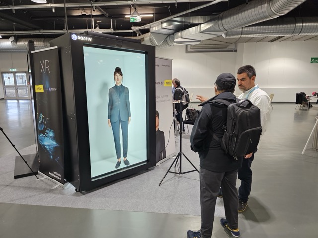 Two people stand in front of a large vertical screen displaying a life-sized image of a woman in a suit. The setting appears to be an indoor tech expo with industrial decor and informational banners.