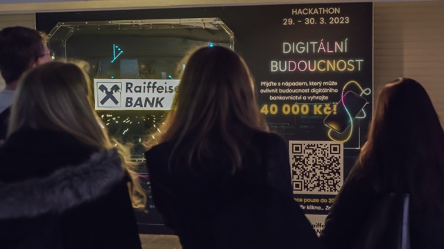 Four people stand facing a brightly lit poster advertising a hackathon event by Raiffeisen Bank with the text “Digitální Budoucnost,” a QR code, prize amount of 40,000 Kč, and dates March 29-30, 2023.