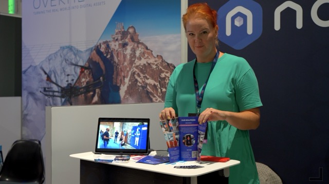 A woman with red hair, wearing a teal dress, stands at a booth holding brochures. A tablet displaying images and more brochures are on the table. A mountain graphic is visible in the background.