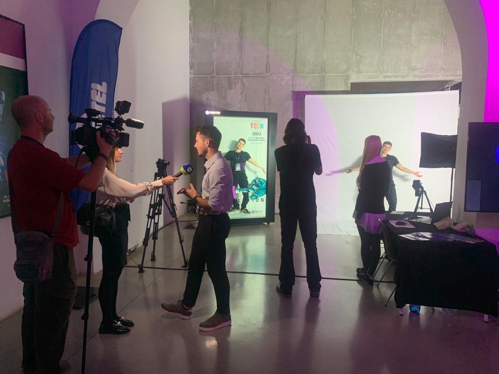 A group of people are at an indoor event. Two are filming an interview with a microphone, while others are near a lit backdrop where a person poses with arms outstretched. Equipment and banners are visible in the room.