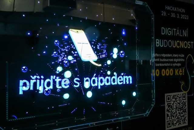 A digital sign displays a glowing smartphone and the Czech text “přijďte s nápadem.” In the background, information about a hackathon and a QR code are visible. The scene is illuminated with blue and white lights.
