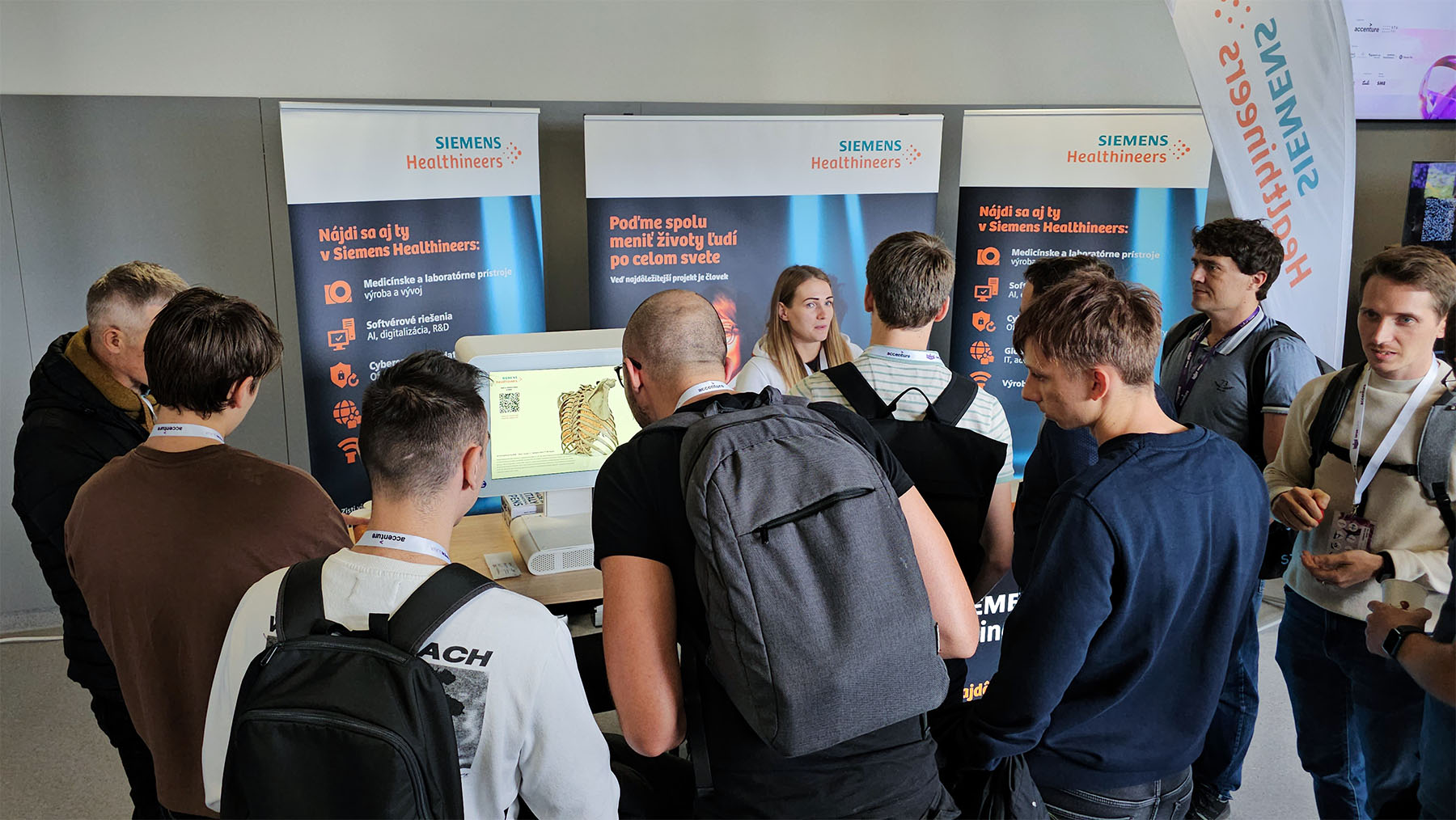 A group of people gather at a Siemens Healthineers booth, watching a presenter explain a medical device shown on a screen.