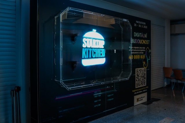 A large illuminated sign reads Startup Kitchen on a dark wall, next to details about a hackathon event and a QR code. Some chairs are visible on the right side of the image.