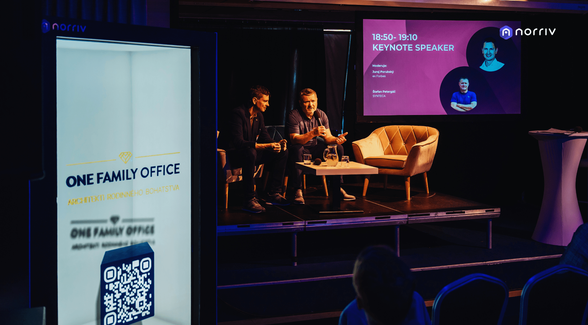 Two men sit and speak on a stage during a keynote session at an event. A screen behind them displays their photos and details. In the foreground, a lit display advertises One Family Office with a QR code.