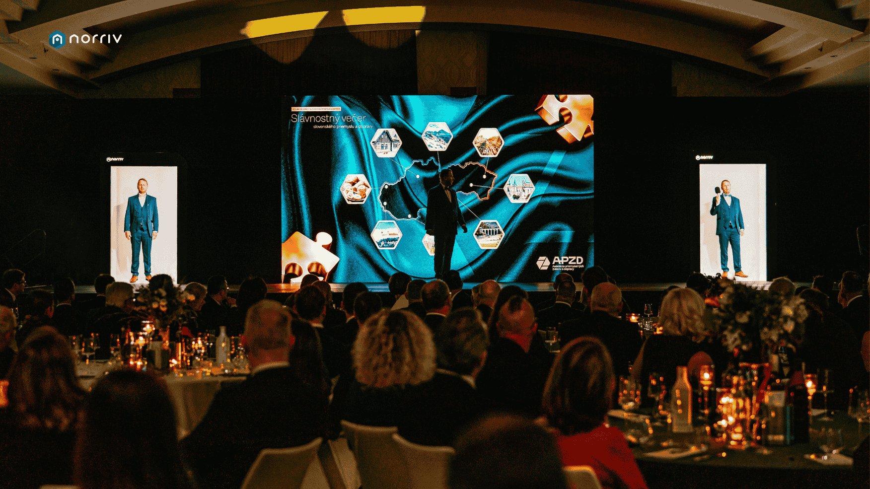 A keynote speaker in a blue suit presents on stage to a seated audience at a formal event, with large digital screens displaying graphics and text behind him. The room is warmly lit with candles on the tables.