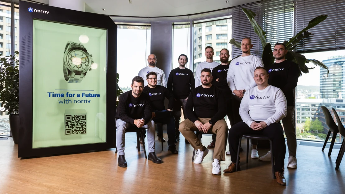 Ten people wearing branded sweatshirts pose in an office with large windows. A digital display shows a wristwatch, the text Time for a Future with norriv, and a QR code. Tall plants and city buildings are visible outside.