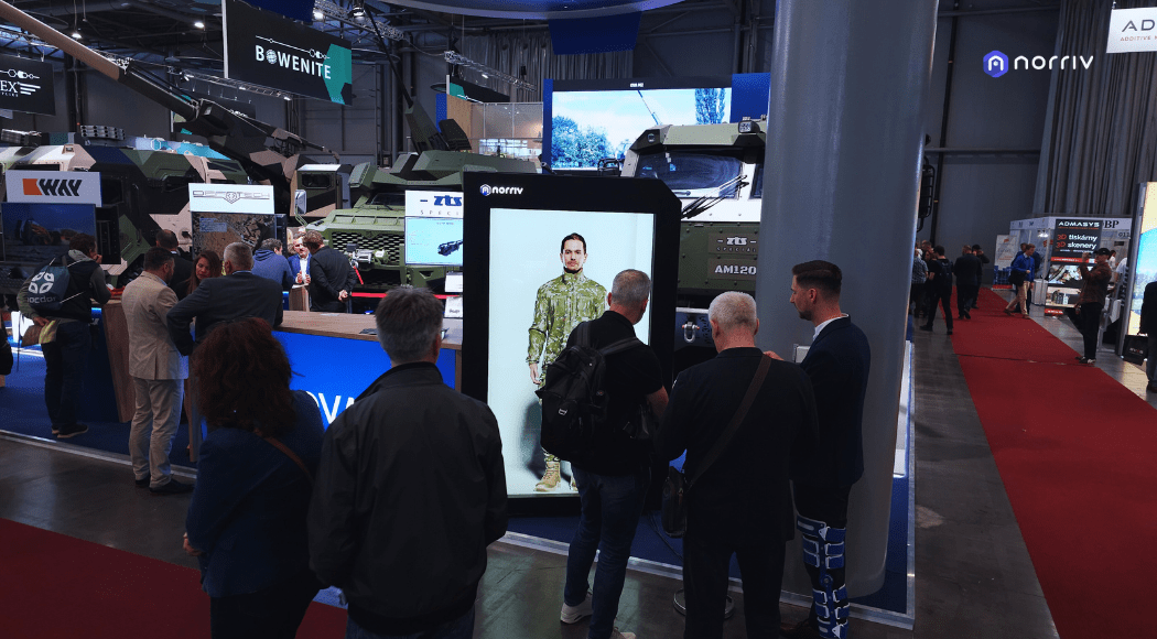 People gather around a digital display showing a person in camouflage clothing at a technology expo, with military vehicles, exhibition booths, and signage visible in the background.