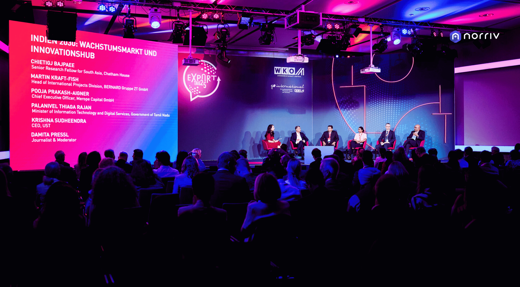 A panel of five speakers sits on stage discussing under pink and purple lighting in front of an audience. Presentation slides and event branding are projected behind them, showing speaker names and event details.