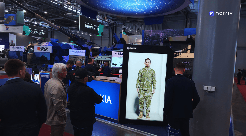 Several people stand around a large digital display showing a man in military camouflage attire at an indoor exhibition with vehicles and technology booths in the background.