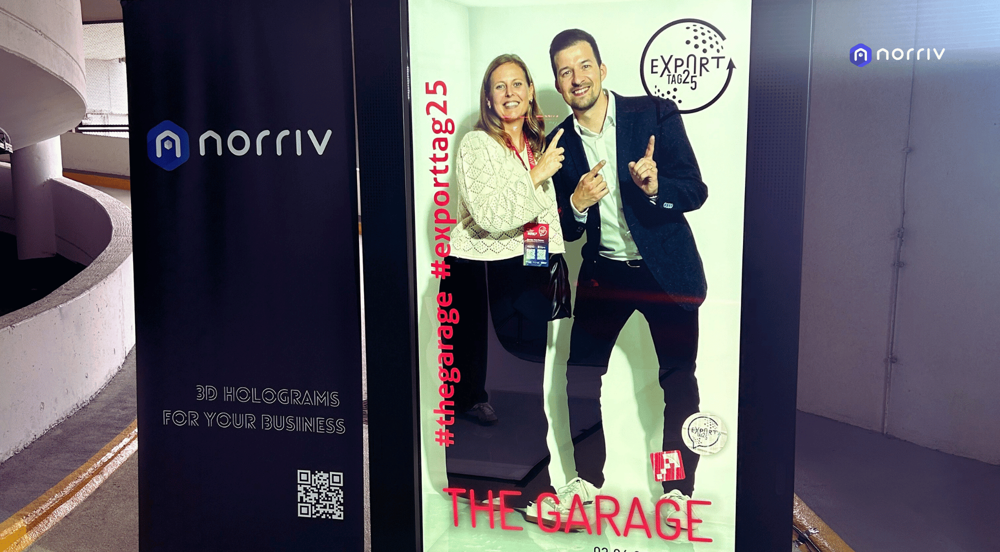 A woman and a man stand smiling and posing inside a display labeled THE GARAGE with #thegarage #exporttag25 and EXpORTTA logos. A nearby banner reads norriv 3D HOLOGRAMS FOR YOUR BUSINESS.