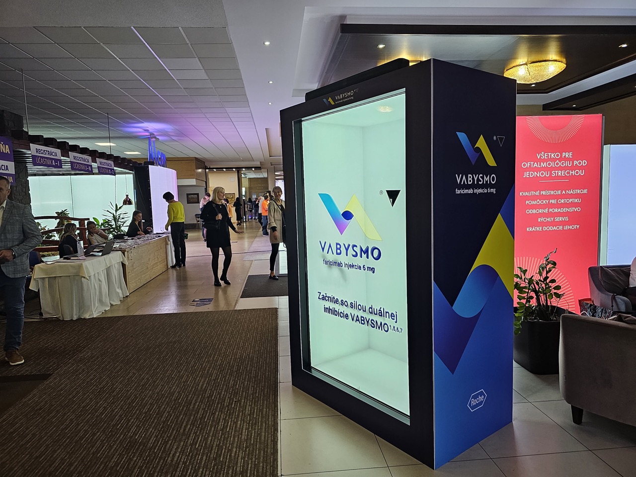A modern indoor event with booths and people walking. The foreground shows a large illuminated display with the Vabysmo logo and text in Slovak. Other booths and attendees are visible in the background.