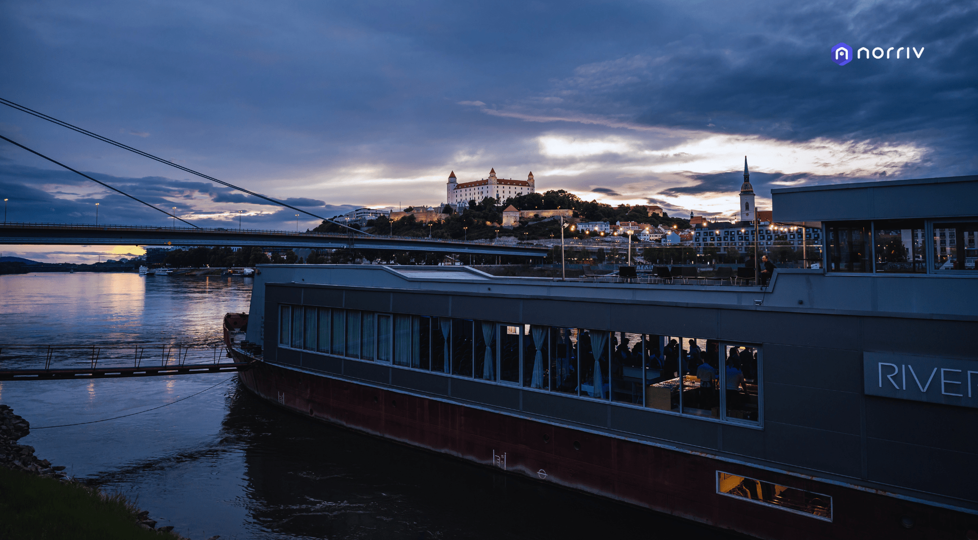 A riverboat docked by the water at dusk, with a bridge spanning across the scene and a hilltop castle illuminated in the background under a cloudy sky.