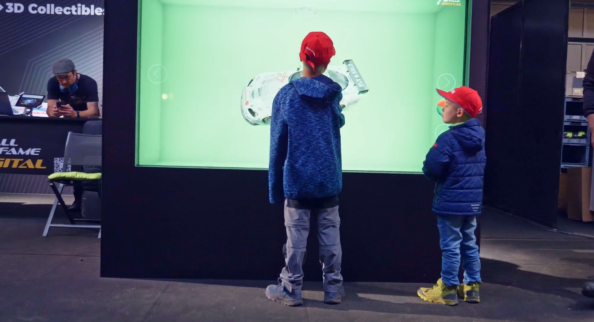 Two children in red caps stand in front of a large interactive display with a green background, observing a 3D object. An adult sits in the background to the left, working at a booth.