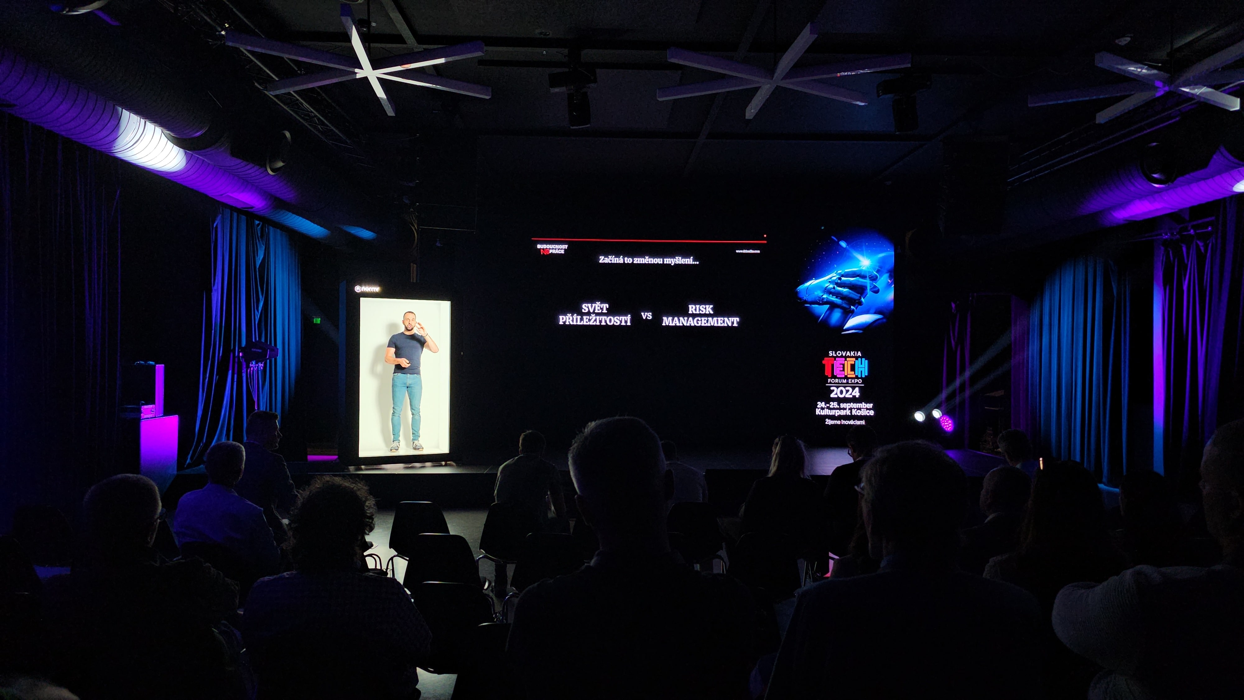 A dark auditorium with people seated, watching a large screen displaying a man on the left and text in the center and right. The room is lit with purple and blue lights, and it appears to be a tech conference or presentation.
