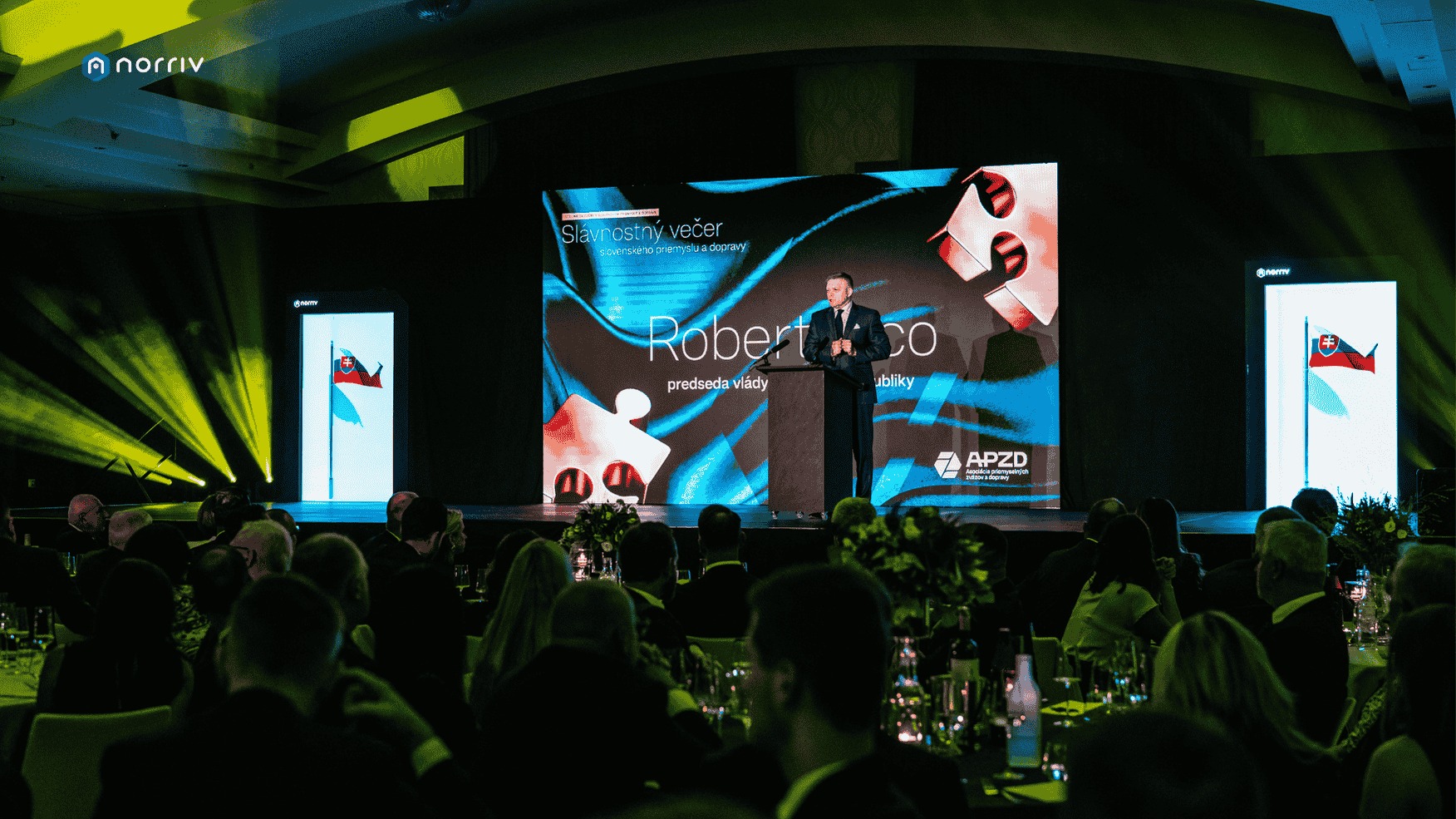 A man in a suit speaks at a podium on a stage during a formal event, with an audience seated at round tables. Large screens and spotlights illuminate the stage, displaying a presentation with colorful graphics.