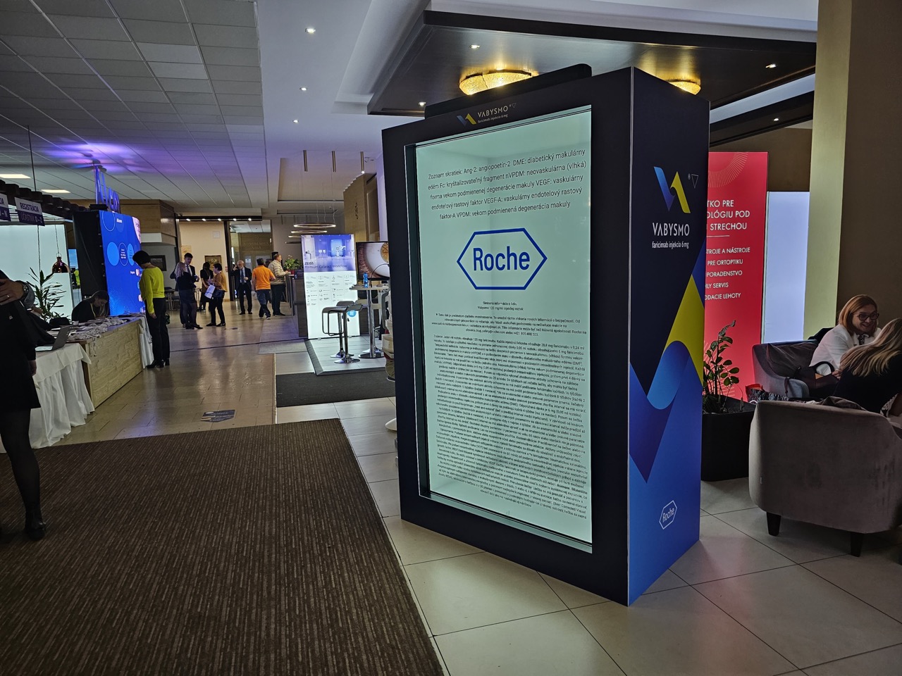 An indoor event space with a large digital display showing Roche’s logo and text. People are gathered near exhibition booths and displays in a brightly lit area with a modern ceiling.