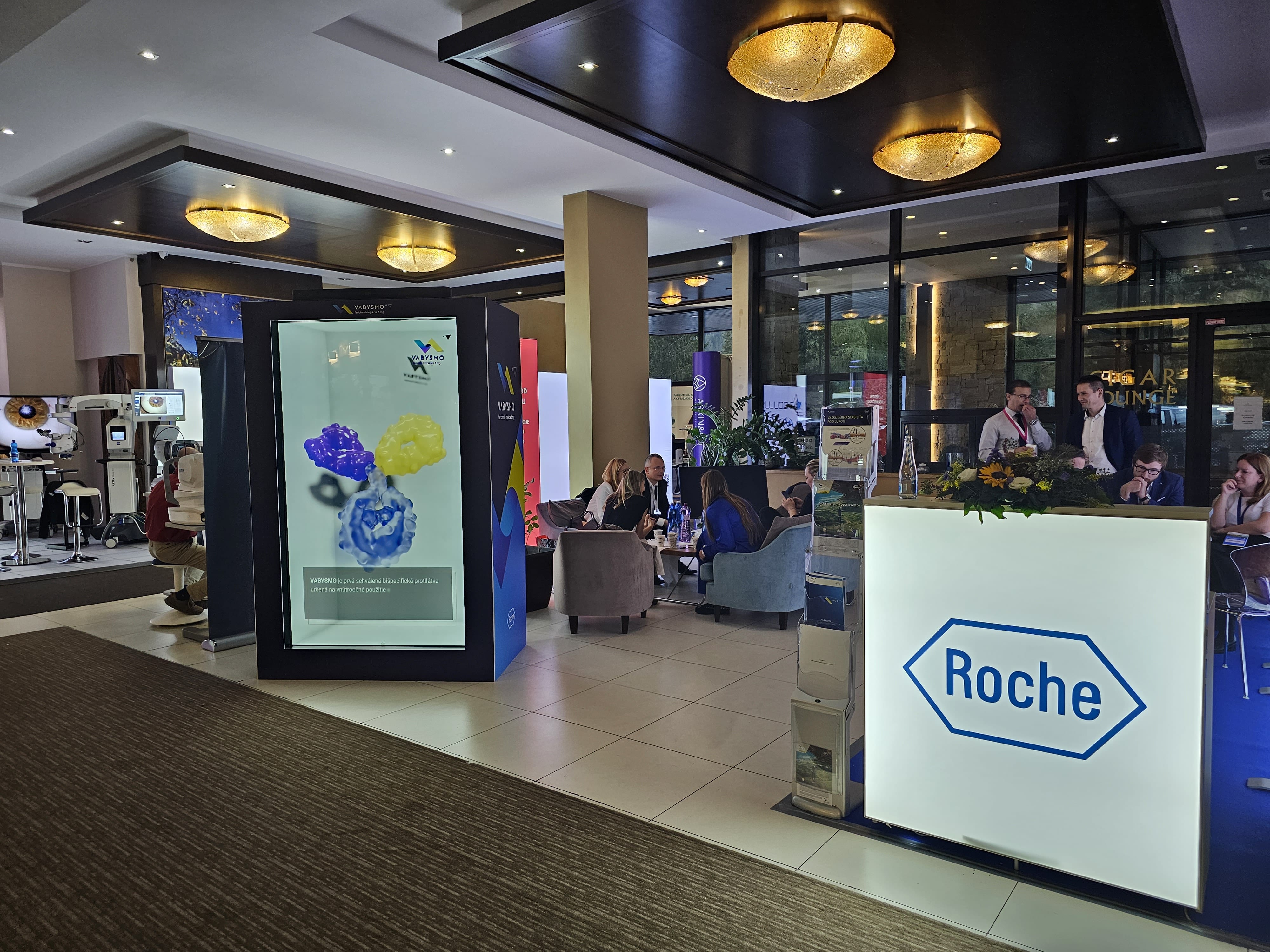 A modern conference lobby features a large illuminated Roche sign at a reception desk, people conversing in seating areas, and a digital display with colorful graphics. The space is brightly lit with ceiling lights.
