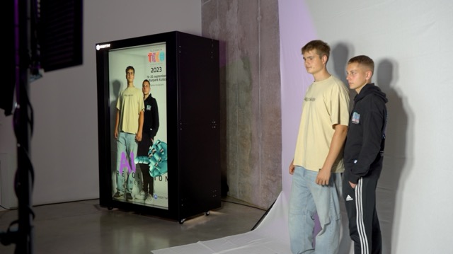 Two young men stand in a studio beside a large digital screen displaying their image and event graphics. The setup includes soft lighting, a white floor mat, and a concrete wall backdrop.