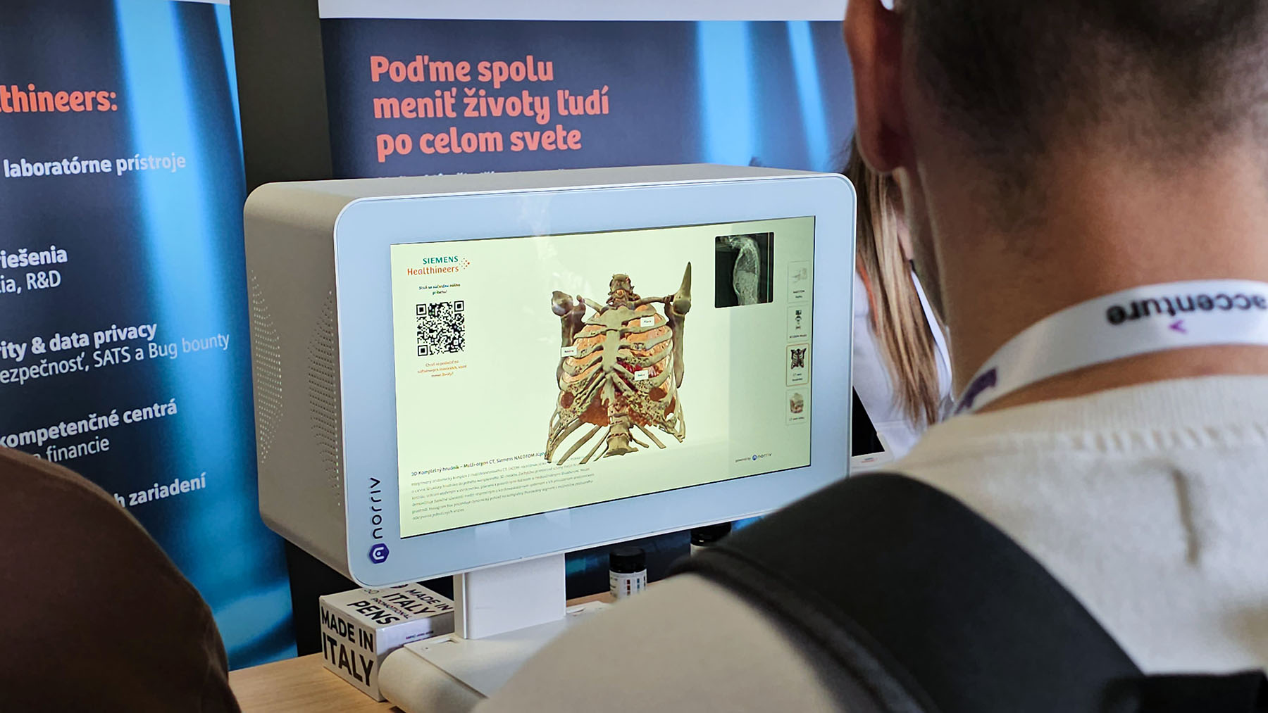 A person examines a digital anatomical model of a human torso on a medical display screen at an exhibition booth.