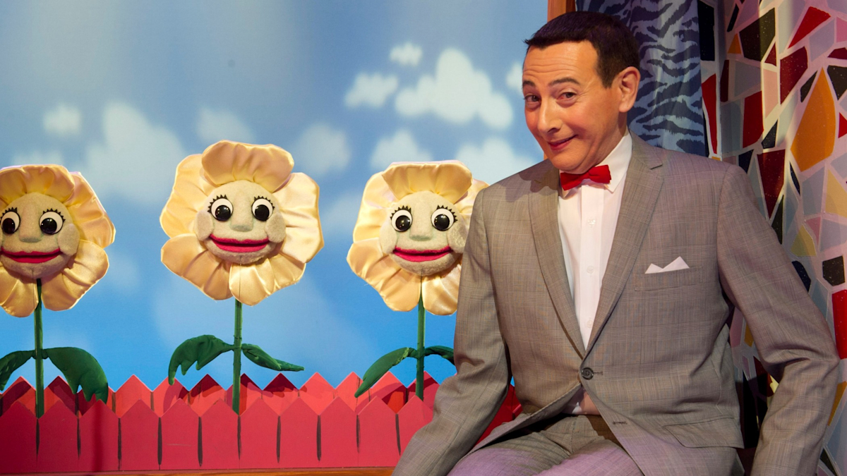 Paul Reubens as Pee-wee Herman smiles while sitting near a red fence with three sunflowers that have faces.