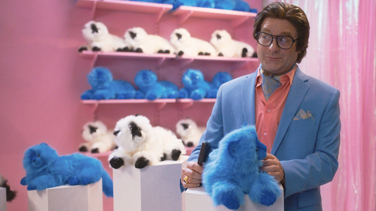 Zach Galifianakis, portraying Ty Warner of Beanie Babies fame, wears a powder-blue suit and holds a fuzzy stuffed animal in a chalky-pink painted room with a pink curtain.