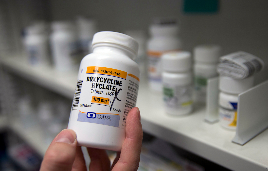 Close-up of a pharmacist's hand holding a bottle of the antibiotic doxycycline hyclate. Other medications in bottles are on shelves in the background of the photo.