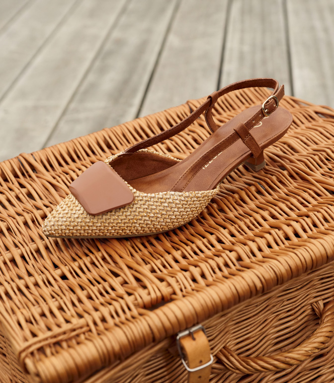 Scarpa slingback con dettagli in rafia beige e pelle marrone su un cesto di vimini su uno sfondo di legno chiaro