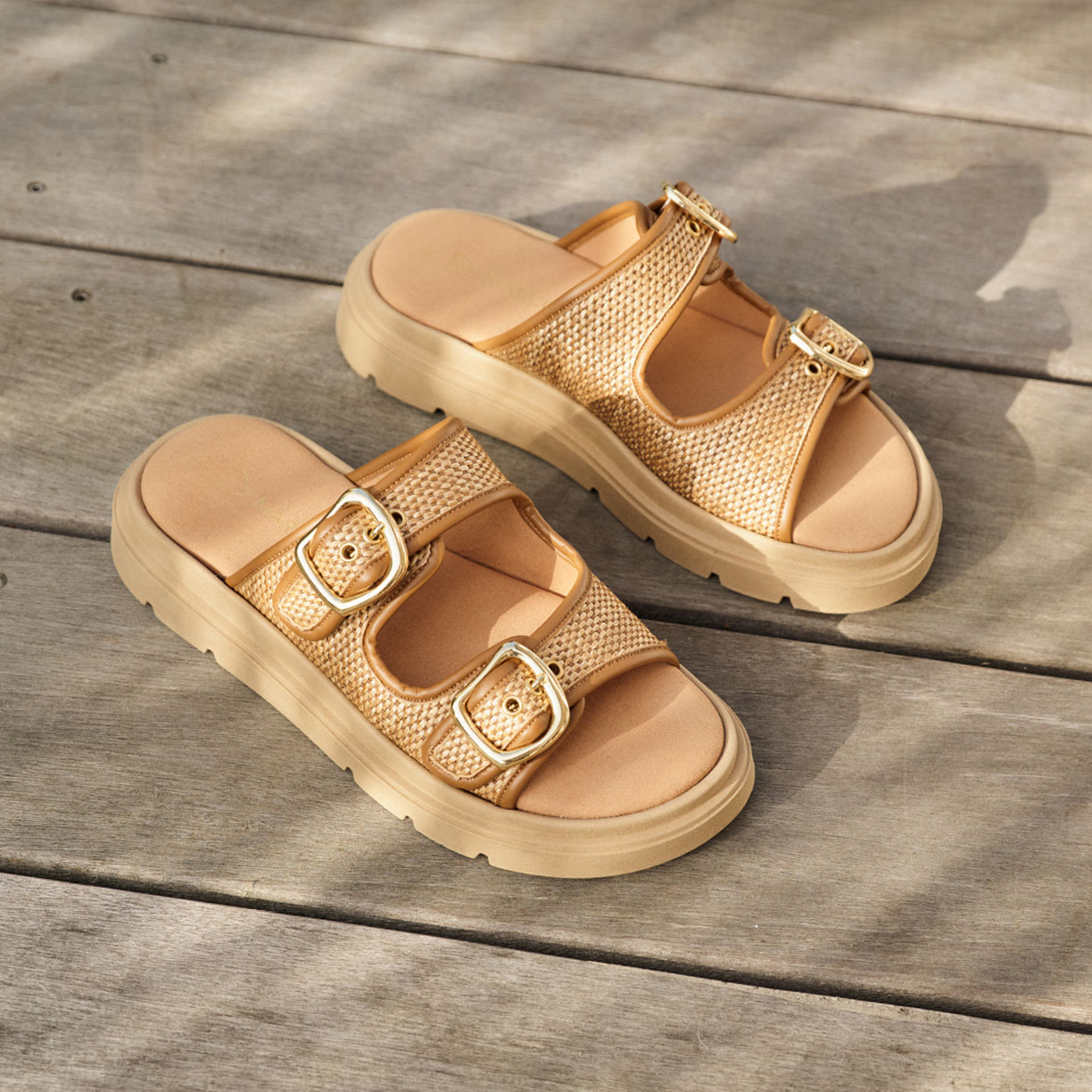 Beige Sandalen mit zwei goldenen Schnallen und Plateausohle auf Holzboden im Sonnenlicht