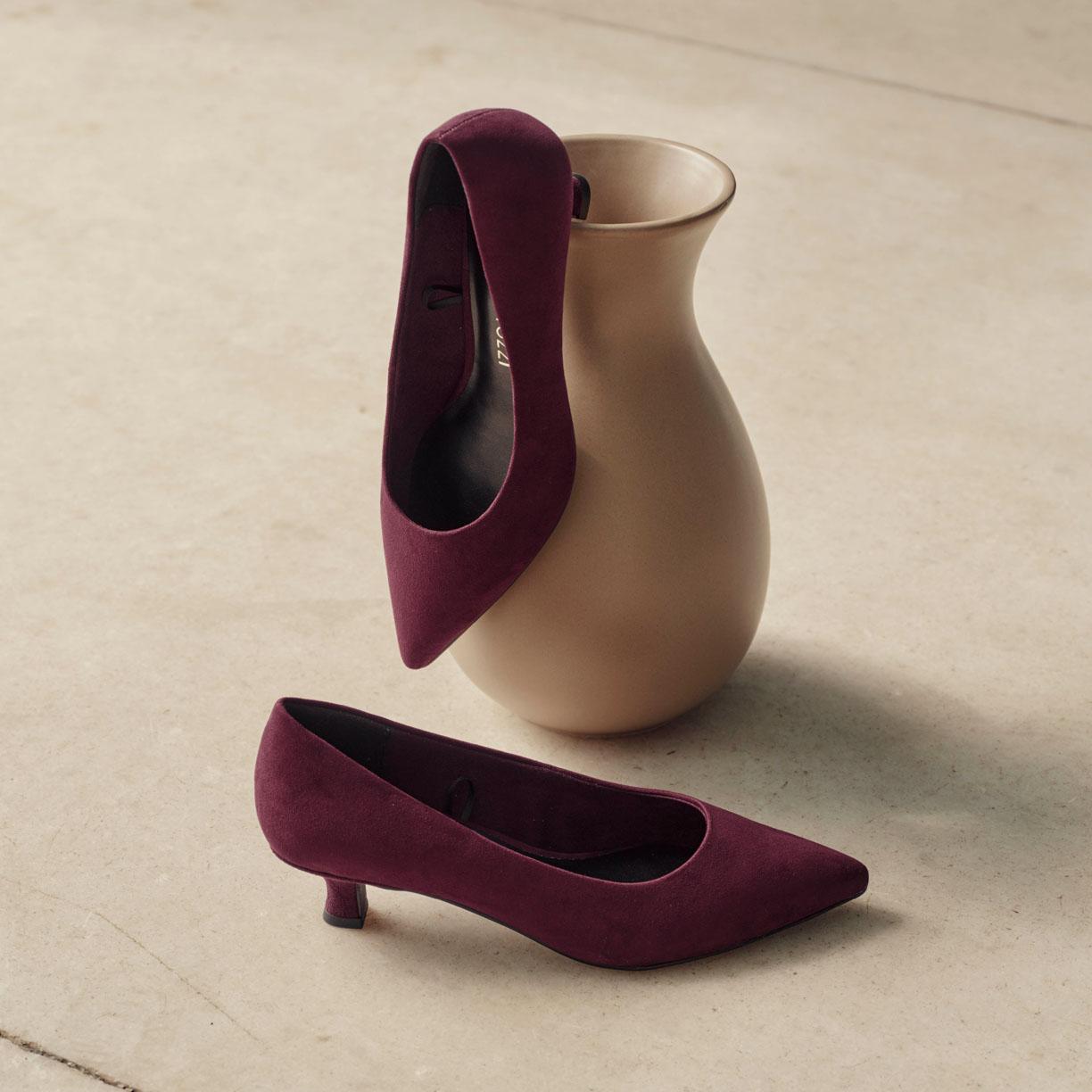 Décolleté con tacco basso color bordeaux e punta affusolata, disposte accanto a un vaso in ceramica beige su uno sfondo chiaro
