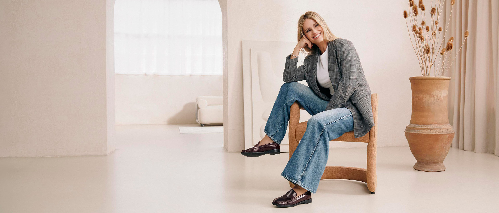 Una donna bionda è seduta sorridente su una poltrona beige in una stanza luminosa; indossa un cappotto a quadri, jeans e mocassini; sullo sfondo si vede un vaso
