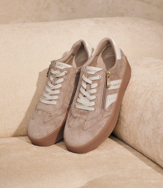 Beige Wildleder-Sneaker mit weißen Schnürsenkeln und seitlichem Reißverschluss auf beigem Sofa in warmem Licht.