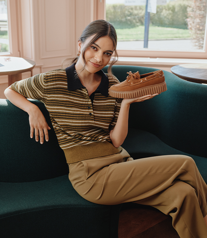 Frau sitzt auf grünem Sofa und präsentiert braunen Wildleder-Schuh, trägt gestreiftes Poloshirt und beige Hose.