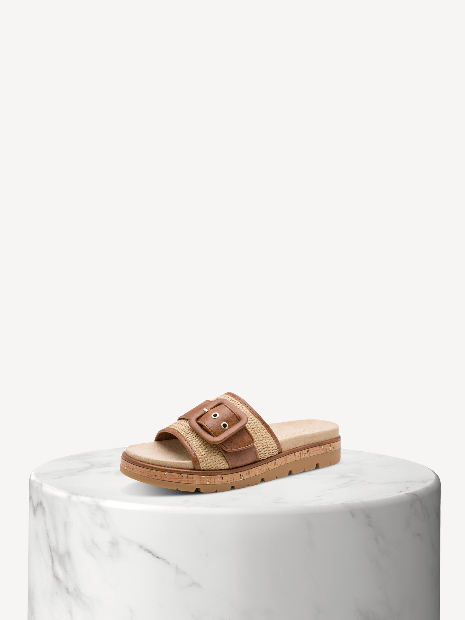 Beige Sandale mit gehäkelten Riemen und goldenen Schnallen auf weißem Marmorsockel vor hellem Hintergrund