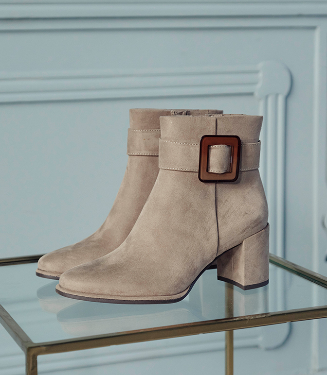 Beige Wildleder-Stiefeletten mit brauner Schnalle und Blockabsatz auf Glastisch vor hellblauer Wand