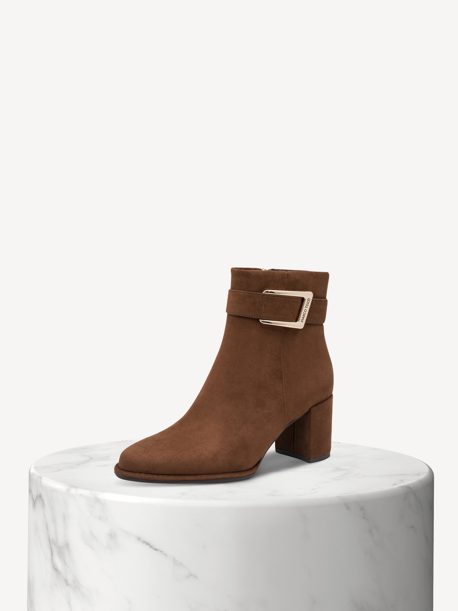 Stivaletti in pelle scamosciata marrone con tacco a blocco e fibbia argentata su base in marmo bianco, su sfondo beige.