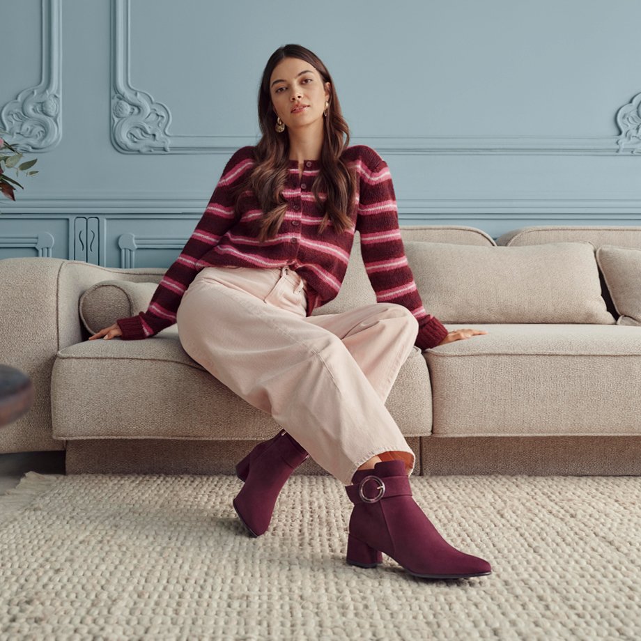 Frau sitzt auf beigem Sofa in gestreiftem bordeauxfarbenem Pullover, heller Hose und bordeauxfarbenen Stiefeln vor hellblauer Wand.