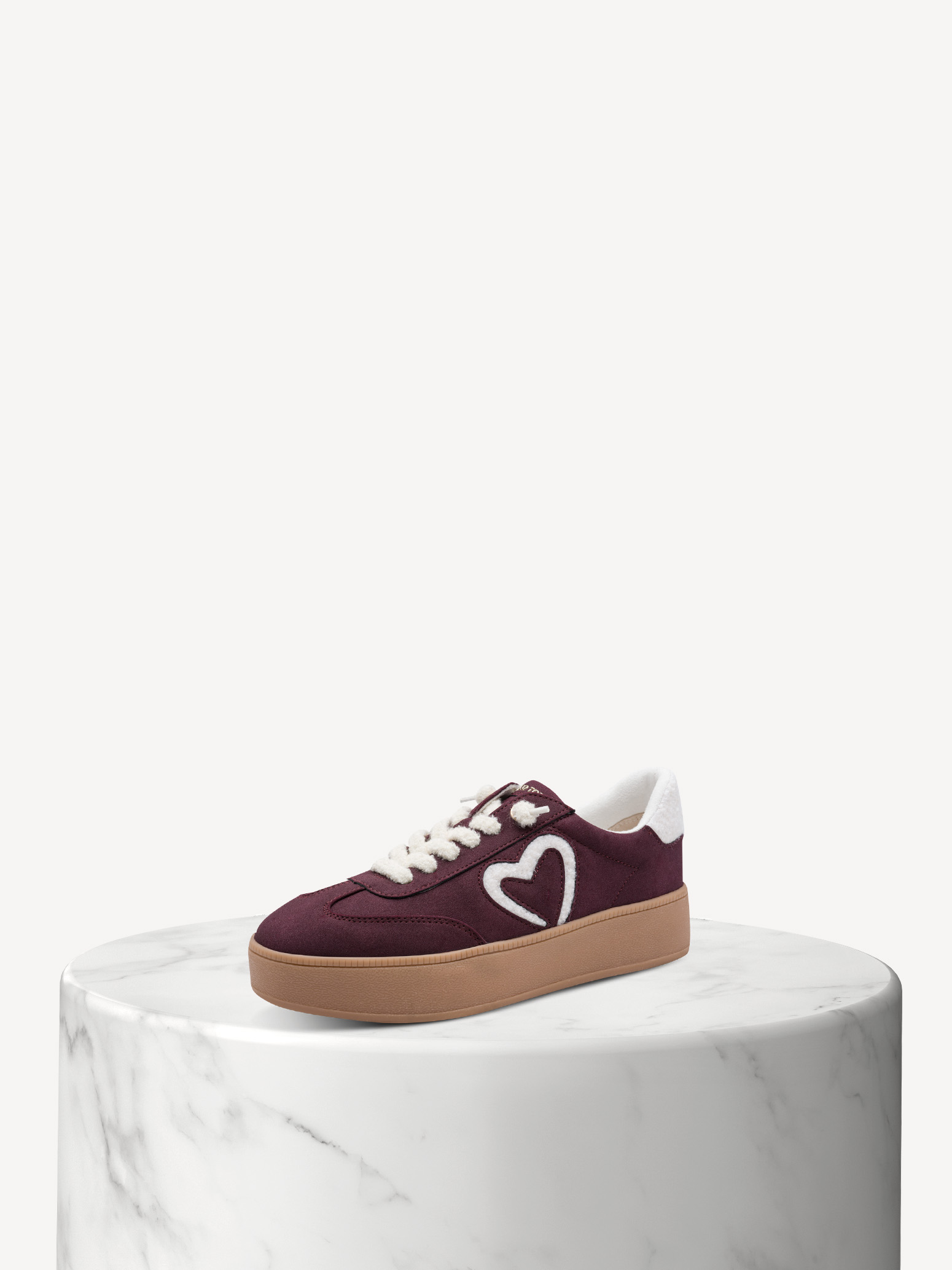 Bordeauxroter Sneaker mit weißem Herz-Logo und beiger Sohle auf weißem Marmorsockel vor hellem Hintergrund.