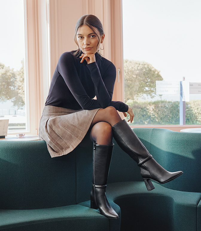 Una donna con un top nero e una gonna beige è seduta su un divano verde; indossa stivali neri, con un interno chiaro sullo sfondo.