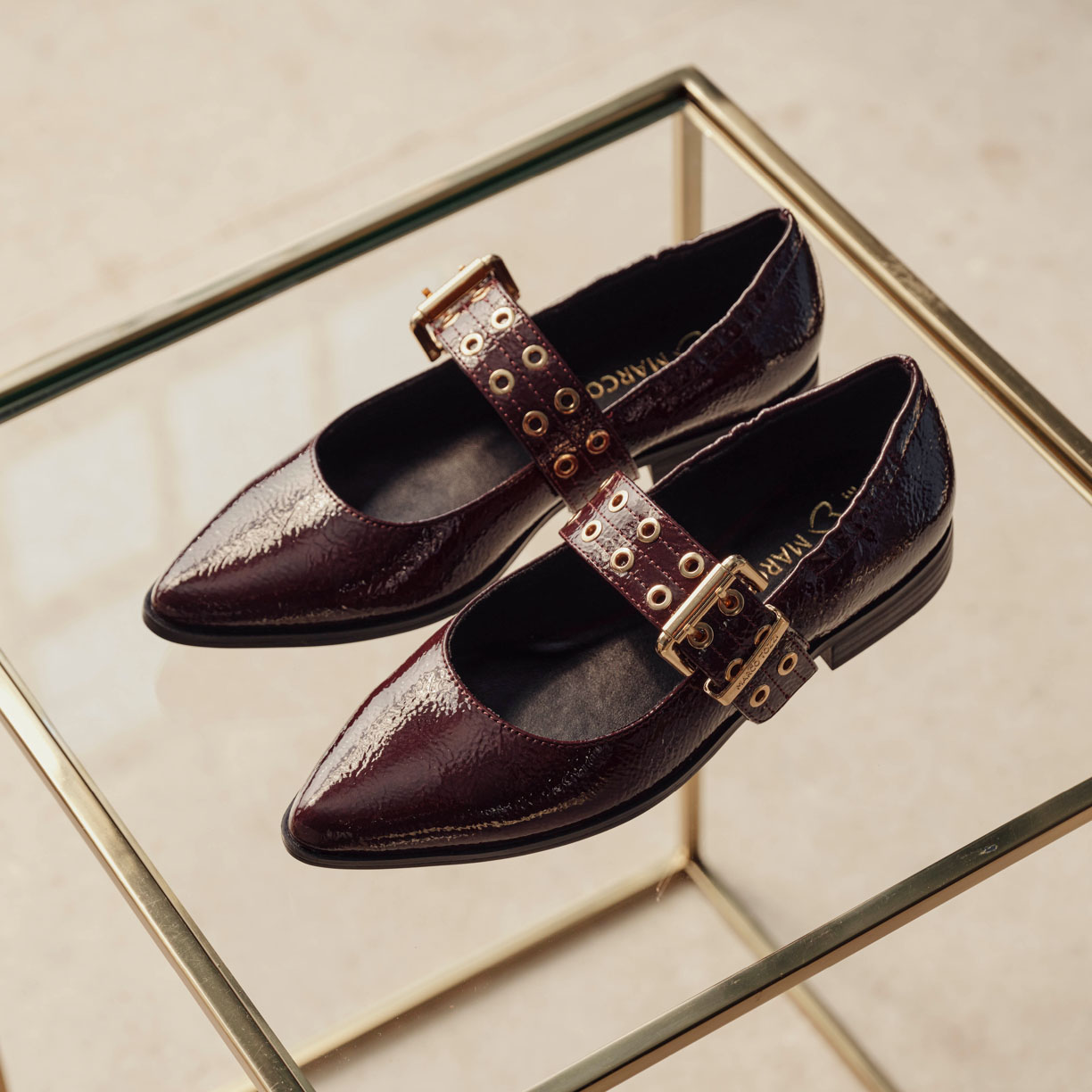 Scarpe in pelle verniciata rosso scuro con fibbia dorata su un vassoio di vetro con cornice dorata su uno sfondo beige.
