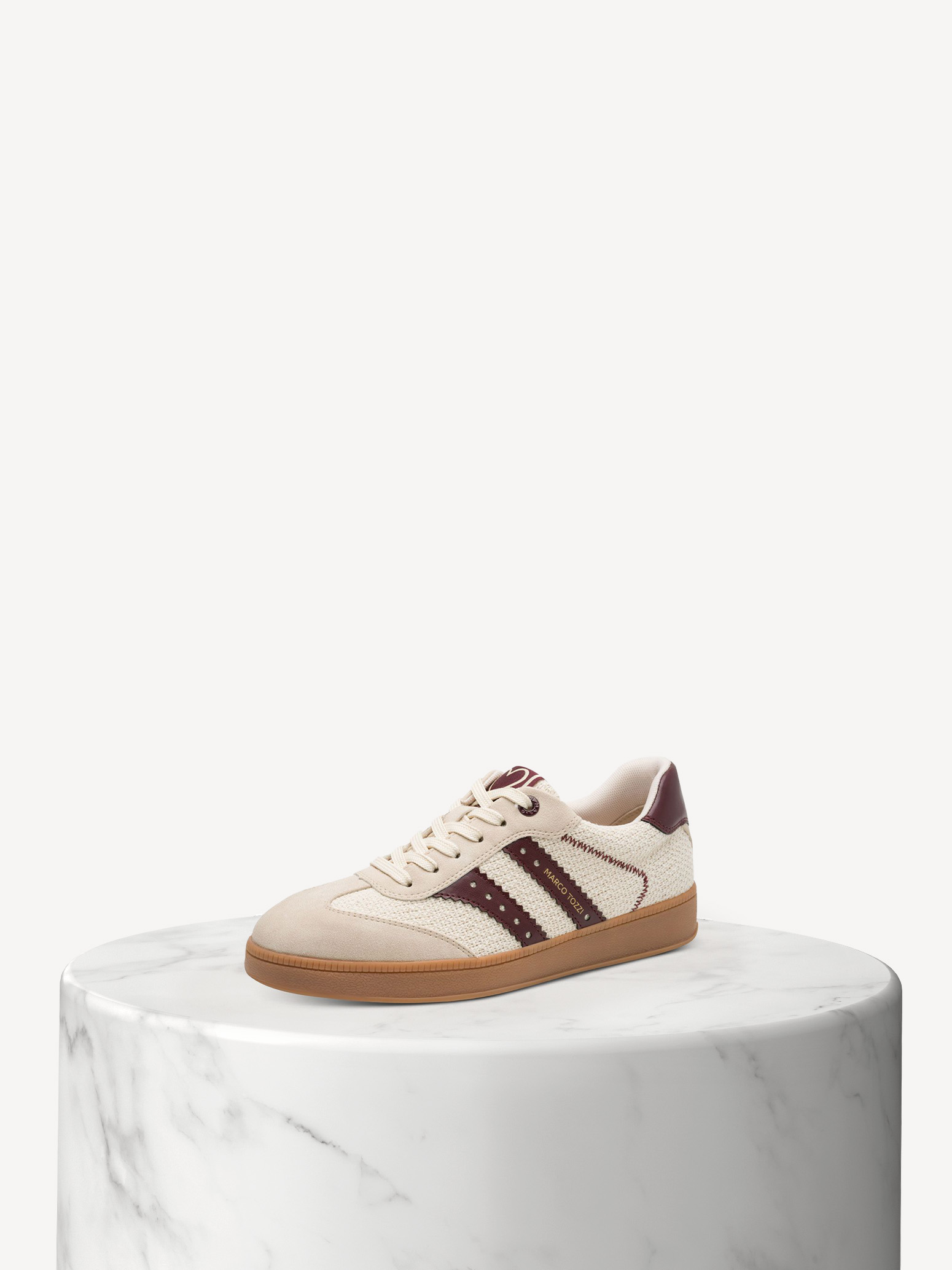 Beiger Sneaker mit bordeauxroten Streifen und Gummisohle auf weißem Marmorsockel vor hellem Hintergrund.