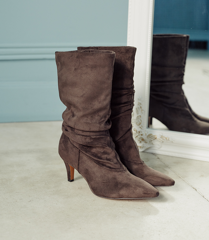 Braune Wildleder-Stiefel mit mittelhohem Absatz und gerafftem Schaft vor einem Spiegel auf hellem Untergrund.