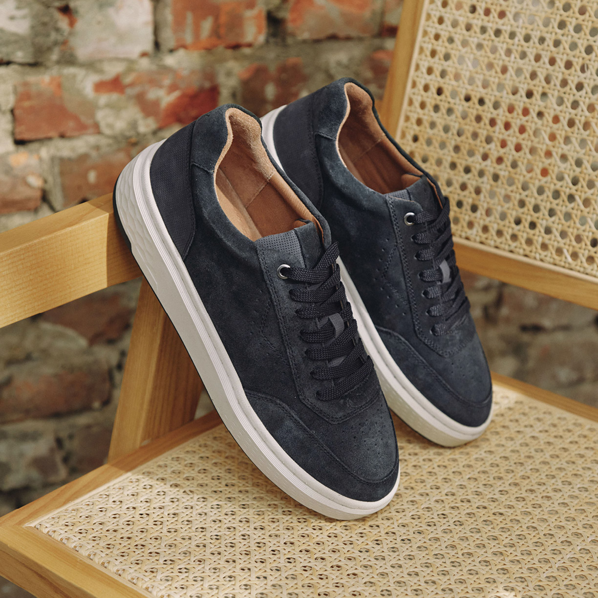 Dunkelblaue Wildleder-Sneaker mit weißer Sohle und braunem Innenfutter auf einem Stuhl mit Rattangeflecht vor Backsteinwand