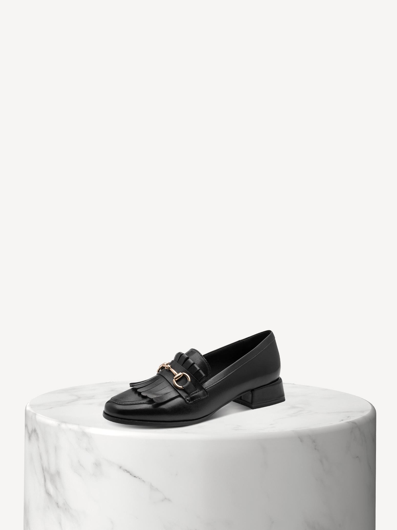 Black leather loafer with fringe and a gold buckle on a white marble base against a beige background.