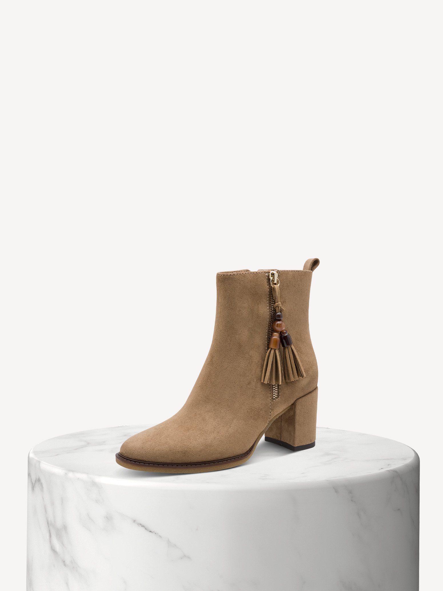 Beiger Wildleder-Stiefelette mit Blockabsatz und Quasten-Reißverschluss auf weißem Marmorsockel vor hellem Hintergrund.