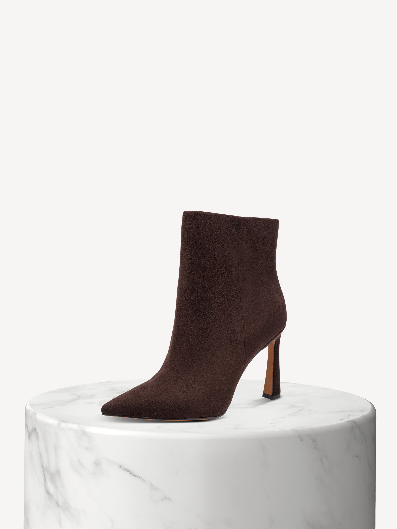 Stivaletti in camoscio marrone scuro con punta affusolata e tacco alto su una base di marmo bianco, su sfondo beige.