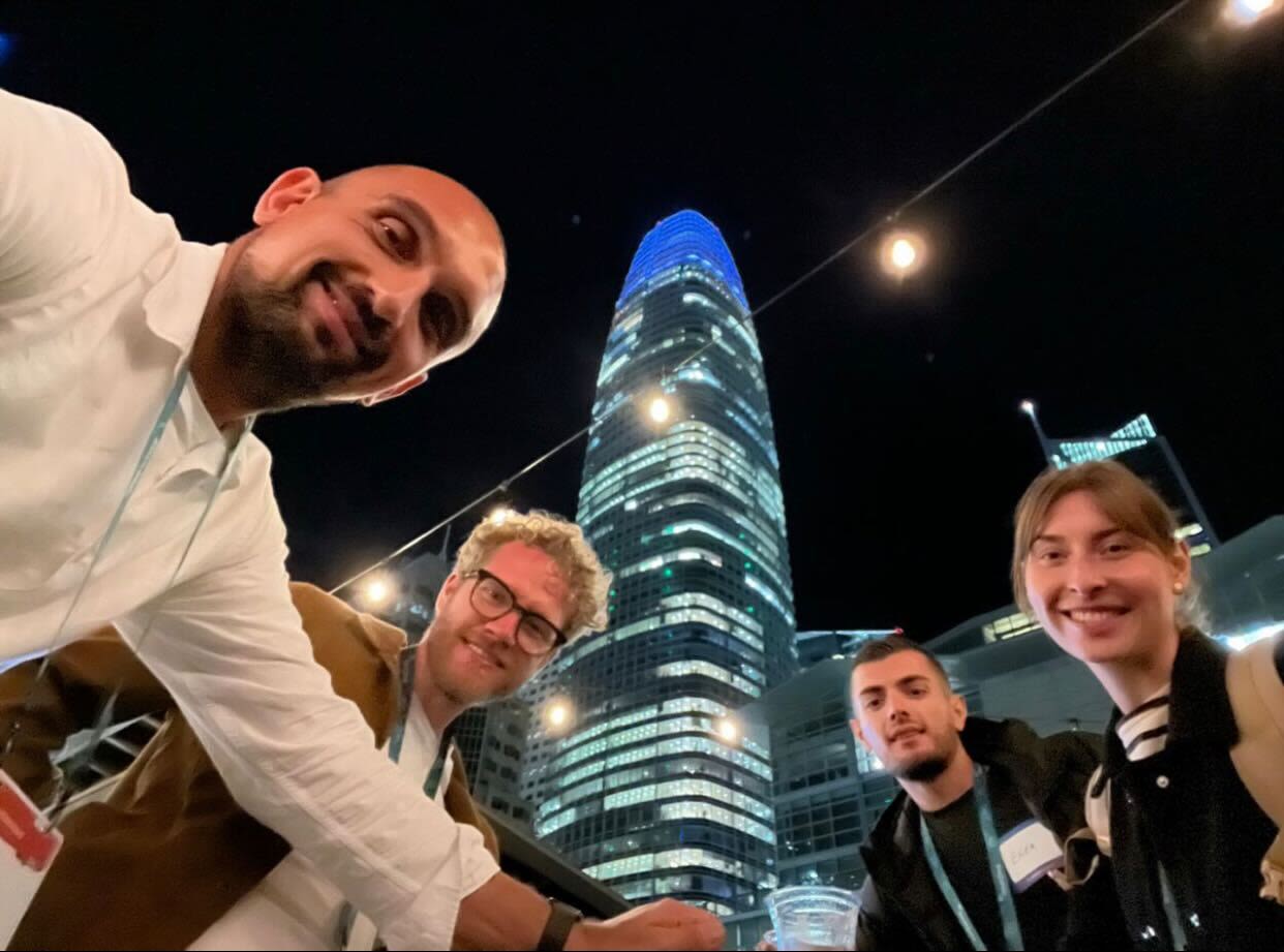 Several person stand in front of a skyscraper one evening and look into the camera