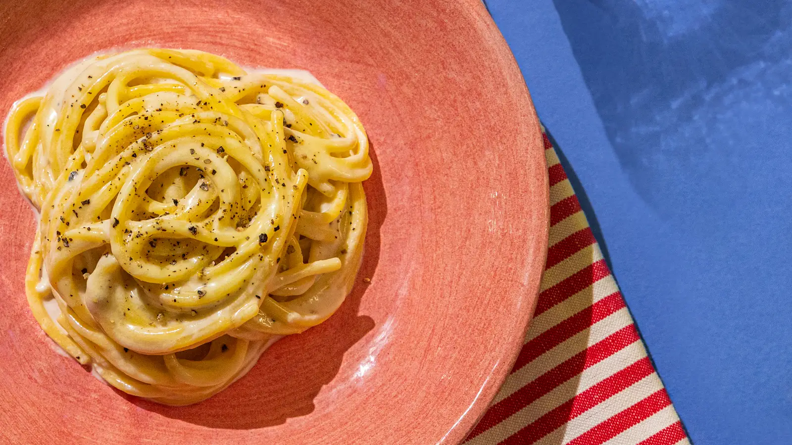 Bowl of pasta with a creamy sauce, on a pink plate sitting on a white, red and blue tablecloth.